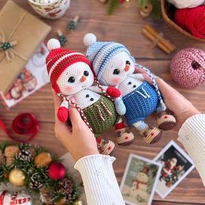 K&ouml;nnte beinhalten: Zwei handgefertigte Schneemann-Puppen aus H&auml;kelarbeit. Ein Schneemann tr&auml;gt eine rot-wei&szlig; gestreifte M&uuml;tze, rote F&auml;ustlinge und eine gr&uuml;ne Latzhose. Der andere tr&auml;gt eine blaue M&uuml;tze, eine blaue Latzhose und rote F&auml;ustlinge. Weihnachten Thema.