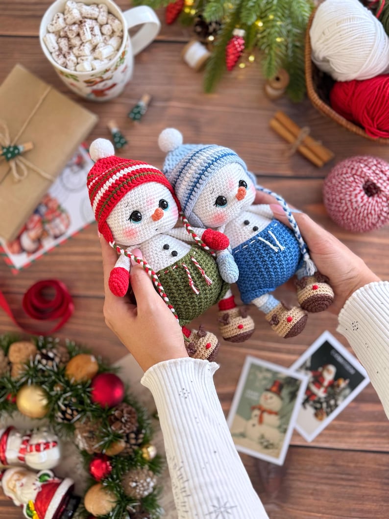 K&ouml;nnte beinhalten: Zwei handgefertigte Schneemann-Puppen aus H&auml;kelarbeit. Ein Schneemann tr&auml;gt eine rot-wei&szlig; gestreifte M&uuml;tze, rote F&auml;ustlinge und eine gr&uuml;ne Latzhose. Der andere tr&auml;gt eine blaue M&uuml;tze, eine blaue Latzhose und rote F&auml;ustlinge. Weihnachten Thema.