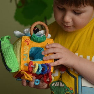 Puede incluir: Un ni&ntilde;o sostiene un juguete de crochet colorido con forma de cubo. El juguete tiene un anillo, una flor y los n&uacute;meros 1, 2 y 3. El juguete est&aacute; hecho de hilo suave y texturizado.