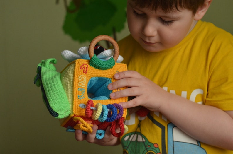 Puede incluir: Un ni&ntilde;o sostiene un juguete de crochet colorido con forma de cubo. El juguete tiene un anillo, una flor y los n&uacute;meros 1, 2 y 3. El juguete est&aacute; hecho de hilo suave y texturizado.