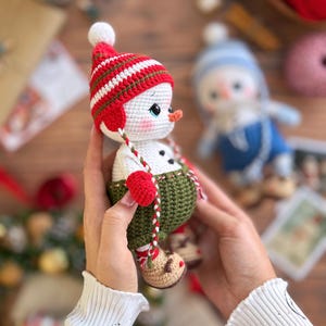 K&ouml;nnte beinhalten: Handgemachte geh&auml;kelte Schneemann-Puppe mit rot-wei&szlig; gestreifter M&uuml;tze, gr&uuml;nen Latzhosen und roten F&auml;ustlingen. Der Schneemann hat eine Karottennase und h&auml;lt eine rot-wei&szlig; gestreifte Schnur. Eine weitere Schneemann-Puppe befindet sich im Hintergrund.