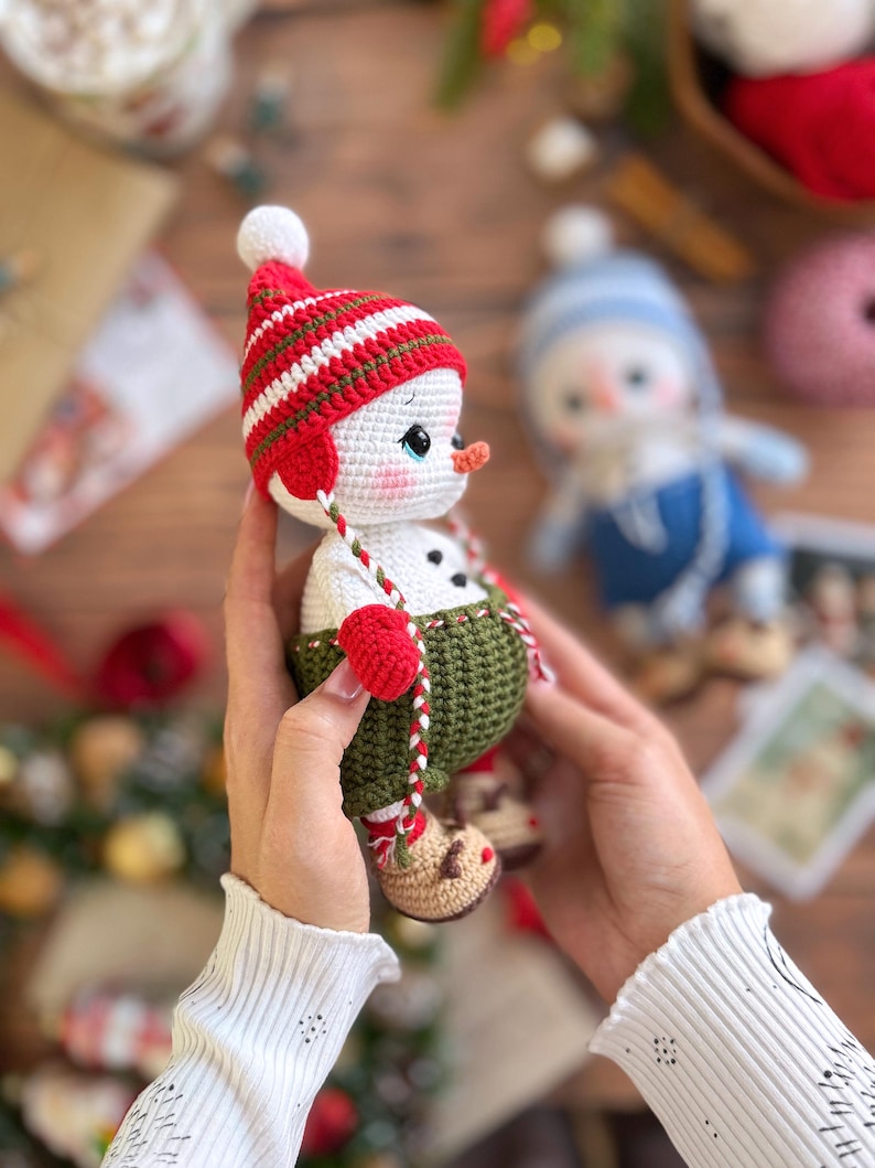 K&ouml;nnte beinhalten: Handgemachte geh&auml;kelte Schneemann-Puppe mit rot-wei&szlig; gestreifter M&uuml;tze, gr&uuml;nen Latzhosen und roten F&auml;ustlingen. Der Schneemann hat eine Karottennase und h&auml;lt eine rot-wei&szlig; gestreifte Schnur. Eine weitere Schneemann-Puppe befindet sich im Hintergrund.