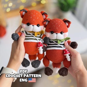 Puede incluir: Dos muñecos de zorro de ganchillo con cuerpos naranjas, caras blancas y camisas a rayas blancas y negras. Cada zorro tiene un pequeño colgante de seta y una bufanda verde o amarilla. El texto "PDF CROCHET PATTERN ENG" es visible.