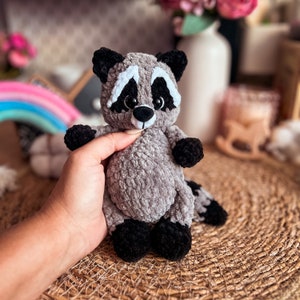Puede incluir: Un juguete de peluche de mapache tejido a crochet con ojos negros, nariz blanca y cuerpo gris y negro. El juguete est&aacute; sentado sobre una superficie de mimbre marr&oacute;n.