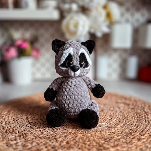 Puede incluir: Un juguete de peluche de mapache gris tejido a crochet con ojos negros y nariz negra. El juguete est&aacute; sentado en una estera tejida marr&oacute;n.