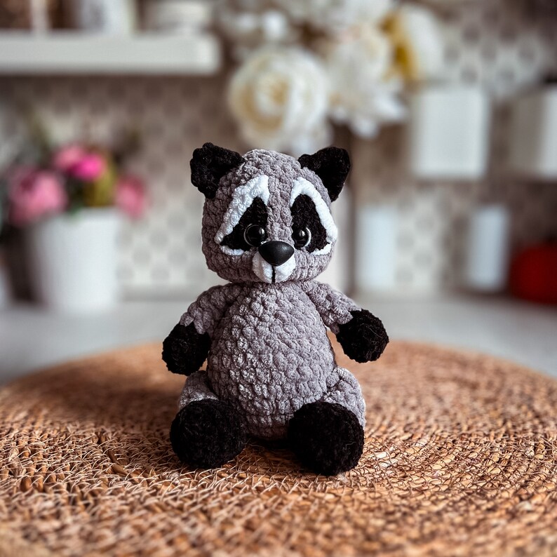 Puede incluir: Un juguete de peluche de mapache gris tejido a crochet con ojos negros y nariz negra. El juguete est&aacute; sentado en una estera tejida marr&oacute;n.