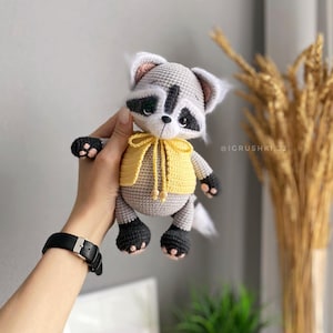 May include: A crocheted raccoon plush toy wearing a yellow hooded sweater. The toy has black eyes, a black nose, and a white tail.