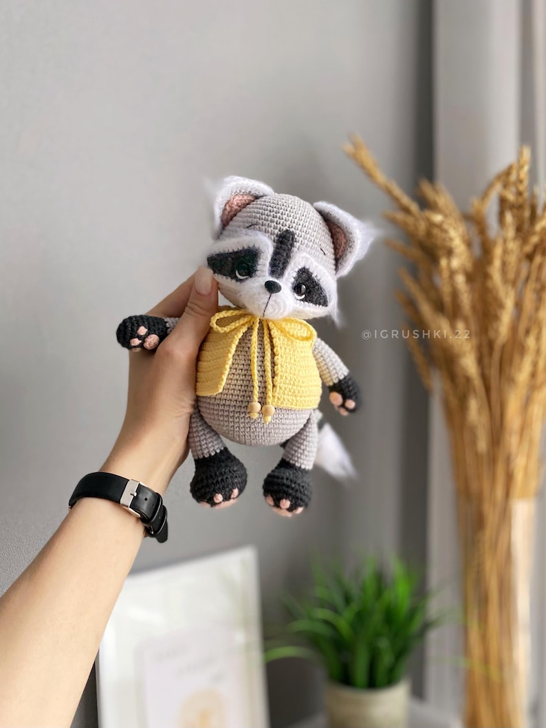 May include: A crocheted raccoon plush toy wearing a yellow hooded sweater. The toy has black eyes, a black nose, and a white tail.