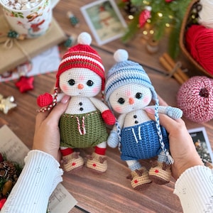 K&ouml;nnte beinhalten: Zwei handgefertigte Schneemann-Puppen aus H&auml;kelarbeit. Der eine Schneemann tr&auml;gt eine rot-wei&szlig; gestreifte M&uuml;tze, gr&uuml;ne Shorts und Rentier-Stiefel. Der andere Schneemann tr&auml;gt eine blau-wei&szlig; gestreifte M&uuml;tze, blaue Shorts und Rentier-Stiefel. Beide Puppen haben schwarze Knopfaugen und Karottennasen.