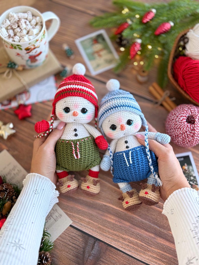 K&ouml;nnte beinhalten: Zwei handgefertigte Schneemann-Puppen aus H&auml;kelarbeit. Der eine Schneemann tr&auml;gt eine rot-wei&szlig; gestreifte M&uuml;tze, gr&uuml;ne Shorts und Rentier-Stiefel. Der andere Schneemann tr&auml;gt eine blau-wei&szlig; gestreifte M&uuml;tze, blaue Shorts und Rentier-Stiefel. Beide Puppen haben schwarze Knopfaugen und Karottennasen.