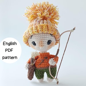 May include: A crocheted doll featuring a large orange pom-pom hat, an orange sweater with a brown bow and arrow symbol, and green pants. The doll holds a brown quiver with arrows and a bow. Text on the left reads: English PDF pattern.