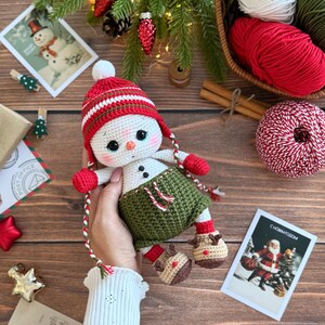 K&ouml;nnte beinhalten: Geh&auml;kelte Schneemann-Puppe mit rot-wei&szlig; gestreifter M&uuml;tze, roten F&auml;ustlingen und gr&uuml;nen Shorts. Der Schneemann hat schwarze Knopfaugen und Stiefel im Rentier-Stil. Weihnachtliche Elemente, einschlie&szlig;lich Garn und Postkarten, sind im Hintergrund.