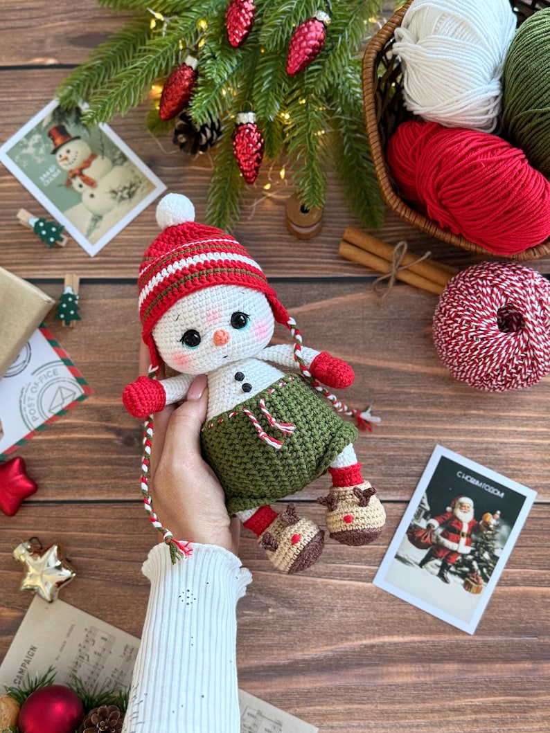 K&ouml;nnte beinhalten: Geh&auml;kelte Schneemann-Puppe mit rot-wei&szlig; gestreifter M&uuml;tze, roten F&auml;ustlingen und gr&uuml;nen Shorts. Der Schneemann hat schwarze Knopfaugen und Stiefel im Rentier-Stil. Weihnachtliche Elemente, einschlie&szlig;lich Garn und Postkarten, sind im Hintergrund.