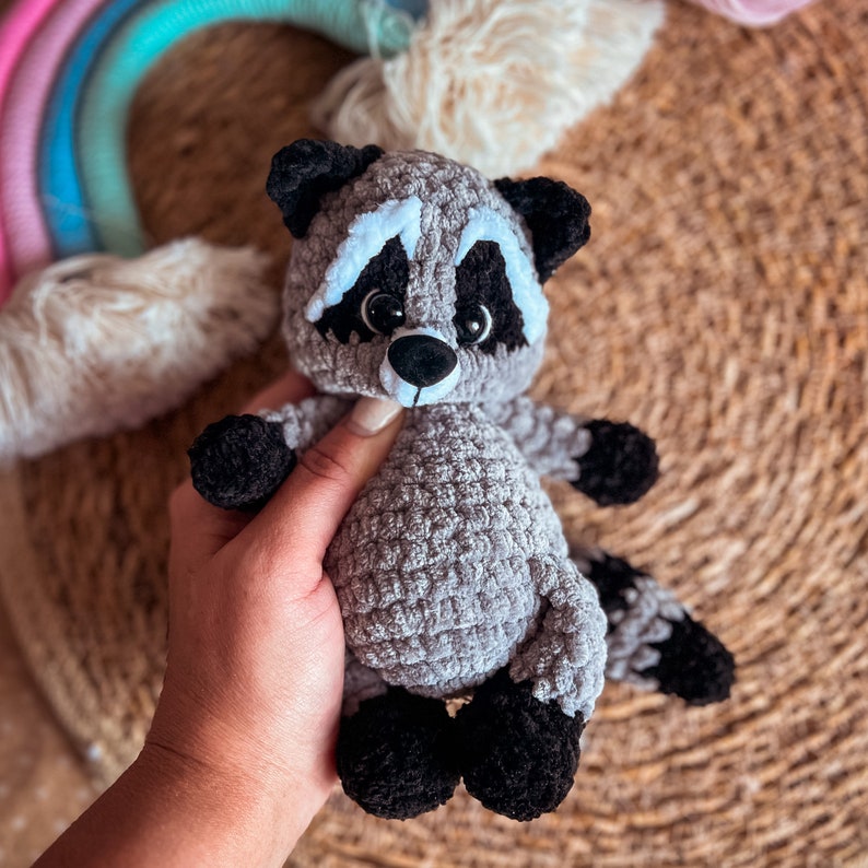 Puede incluir: Un juguete de peluche de mapache de crochet gris con ojos negros y una nariz blanca. El juguete est&aacute; sentado sobre una superficie de mimbre marr&oacute;n.