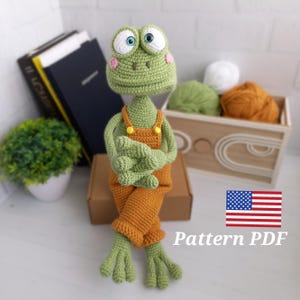 May include: Crochet pattern for a green frog wearing orange dungarees with yellow buttons. The frog has large, white eyes and pink cheeks. The pattern is available as a PDF download. The image also includes a US flag.
