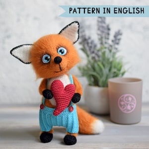 Puede incluir: Un juguete de zorro tejido a ganchillo con pelaje naranja, grandes ojos azules y orejas con puntas negras. Lleva un peto azul claro con botones rojos y sostiene un corazón rojo texturizado. Arriba aparece el texto PATTERN IN ENGLISH.