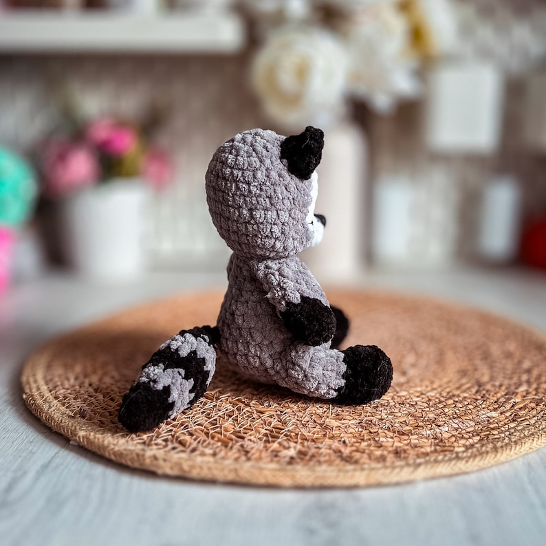 Puede incluir: Un juguete de peluche de mapache tejido a crochet en color gris y negro. El juguete est&aacute; sentado en una estera tejida de color marr&oacute;n.