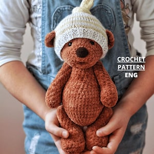May include: A brown crocheted teddy bear wearing a cream-colored hat with a yellow stripe. The bear has a dark brown nose and is held in someone's hands. The text "CROCHET PATTERN ENG" is visible on the right side.