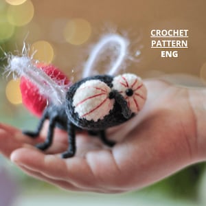 Può includere: Una mosca all'uncinetto con grandi occhi dettagliati e dettagli rossi e bianchi. La mosca ha un corpo, zampe e antenne neri e ali rosse. Il testo "CROCHET PATTERN ENG" è visibile.