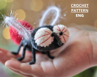 May include: A crocheted fly with large, detailed eyes and red and white accents. The fly has a black body, legs, and antennae, and red wings. The text "CROCHET PATTERN ENG" is visible in the upper right corner.