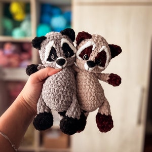 Puede incluir: Dos peluches de mapache tejidos a crochet, uno gris y otro marr&oacute;n, con ojos negros y detalles blancos. El mapache gris est&aacute; sosteniendo al mapache marr&oacute;n en sus brazos.