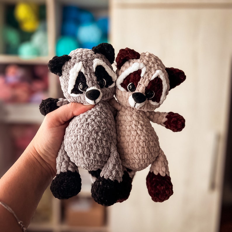 Puede incluir: Dos peluches de mapache tejidos a crochet, uno gris y otro marr&oacute;n, con ojos negros y detalles blancos. El mapache gris est&aacute; sosteniendo al mapache marr&oacute;n en sus brazos.