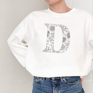 May include: A white sweatshirt featuring a large letter "D" in a floral pattern. The floral design is in shades of gray and white. The sweatshirt has long sleeves and a crew neck.