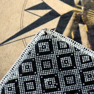 May include: Close-up of a black and white beaded purse corner with a geometric pattern. The purse has a white stitched border. The background features a compass rose design on a tan surface.