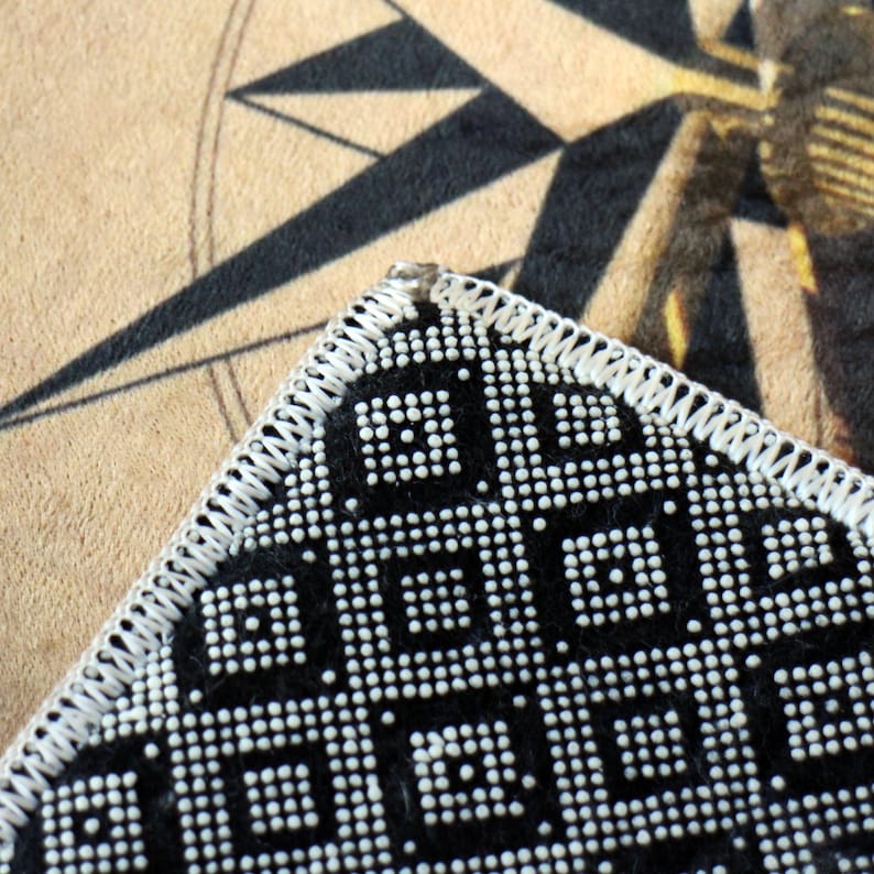 May include: Close-up of a black and white beaded purse corner with a geometric pattern. The purse has a white stitched border. The background features a compass rose design on a tan surface.