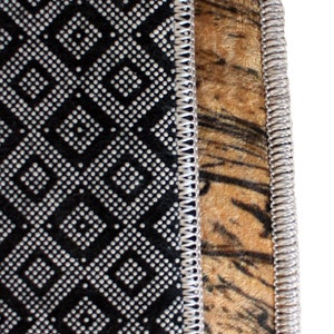 May include: Close-up of a rug with a black and white geometric pattern on the left and a tiger stripe pattern on the right. The edges are trimmed with a silver-colored, stitched border. The rug has a soft, plush texture.