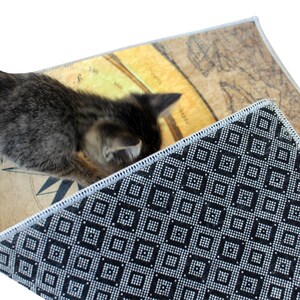 May include: A small, gray tabby kitten explores a rug with a vintage map design. The rug has a cream-colored border and a black and white geometric pattern on the underside. The map design features tan and brown tones.