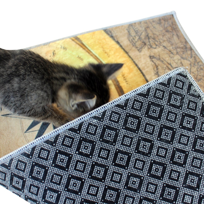May include: A small, gray tabby kitten explores a rug with a vintage map design. The rug has a cream-colored border and a black and white geometric pattern on the underside. The map design features tan and brown tones.