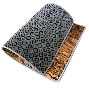 May include: A rectangular mat with a black and white geometric pattern on one side and a brown, wood-grain pattern on the other. The edges are finished with a silver trim. The mat is partially rolled up, revealing both sides.
