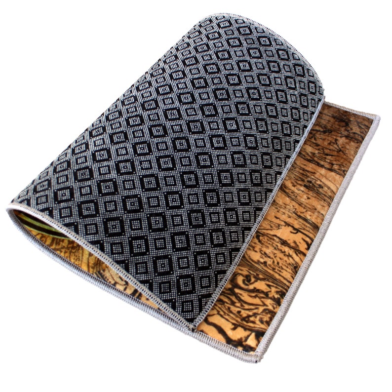 May include: A rectangular mat with a black and white geometric pattern on one side and a brown, wood-grain pattern on the other. The edges are finished with a silver trim. The mat is partially rolled up, revealing both sides.