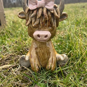 May include: A ceramic Highland cow figurine with a light brown body, a pink bow, and a pink nose. The cow is sitting in a grassy area. The figurine has detailed horns and fur.