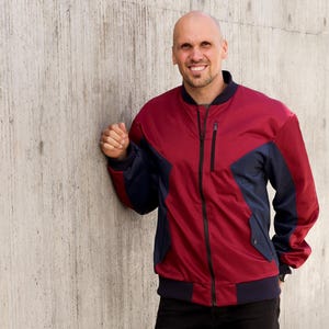 Op de afbeelding: Een rode en marineblauwe bomberjack met een zwarte rits en zakken. De jas heeft een geribbelde kraag en manchetten. De persoon op de foto draagt een zwarte broek.