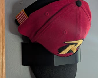 May include: A red baseball cap with a black brim and a yellow 'R' logo. The cap is displayed on a black wall mount.