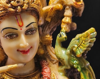 Puede incluir: Una figura detallada de una deidad hindú, probablemente Radha o Lakshmi, adornada con joyas doradas y rojas, un tocado de plumas de pavo real y un pavo real verde posado en su hombro.