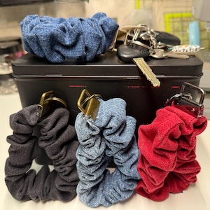 May include: A collection of scrunchies in denim blue, black, and red. Each scrunchie has a metal clasp. One denim blue scrunchie sits atop a black metal box. Keys are on the box. The scrunchies are made of fabric.