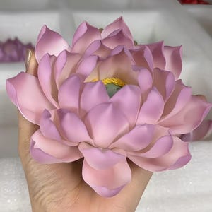 May include: A large, lifelike lotus flower in shades of pink and lavender, held in a hand. The flower's petals are detailed with a gradient of colors, and the center features yellow and green accents. The overall aesthetic is soft and delicate.