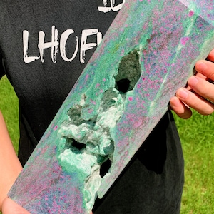 May include: A large, polished crystal with a vibrant mix of green, pink, and blue hues. The crystal has a rough, textured area with a dark cavity. The person in the image is wearing a black t-shirt with the text "TEN LHOF" in white.