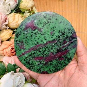 Könnte beinhalten: Ein runder, polierter Zoisitstein mit einem lebendigen grünen Grundton, übersät mit schwarzen Punkten und durchzogen von tiefroten Adern. Der Stein wird in einer Hand gehalten und zeigt seine natürliche Schönheit und einzigartigen Muster. Ideal für die Inneneinrichtung.