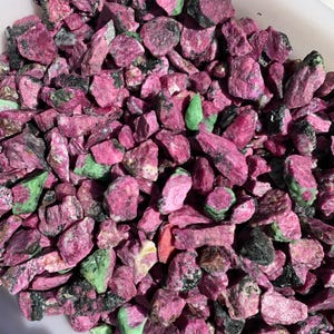 May include: A close-up view of a pile of rough, pink and green gemstones. The stones are various sizes and shapes, with some having a black, speckled appearance.