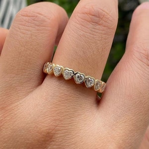 K&ouml;nnte beinhalten: Ein Goldring mit einer Reihe herzf&ouml;rmiger Diamanten. Der Ring befindet sich an einem Finger.