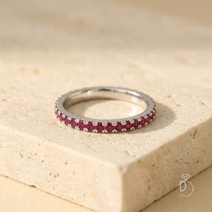 Peut inclure: Une bague alliance en métal argenté ornée d'une rangée continue de petites pierres précieuses rondes d'un rouge vif, serties dans une monture à griffes partagées. L'anneau métallique présente une finition polie et les pierres sont uniformes.