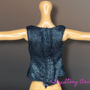 Puede incluir: Una blusa sin mangas de color azul oscuro con una tela texturizada y brillante. La blusa se muestra en un maniqu&iacute; con los brazos extendidos. La parte inferior de la blusa tiene un ligero volante.