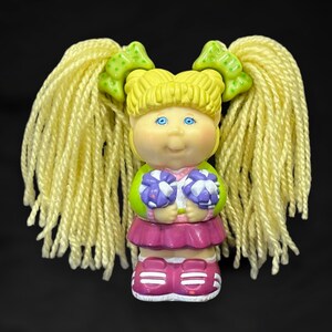 Puede incluir: Una muñeca Cabbage Patch Kid con pelo rubio de lana en coletas, vestida con un uniforme de animadora verde y rosa y sosteniendo pompones morados y blancos.