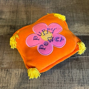 Op de afbeelding: Oranje pluche kussen met een roze bloem geborduurd erop. De bloem heeft een geel centrum en de woorden "Puppy Power" zijn in paars geborduurd op de bloem.