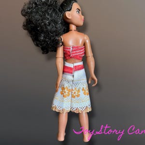 May include: A doll wearing a red and white top and a white skirt with orange floral patterns. The doll has black curly hair and is standing against a brown background.