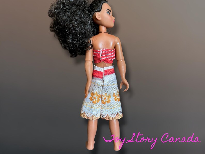 May include: A doll wearing a red and white top and a white skirt with orange floral patterns. The doll has black curly hair and is standing against a brown background.